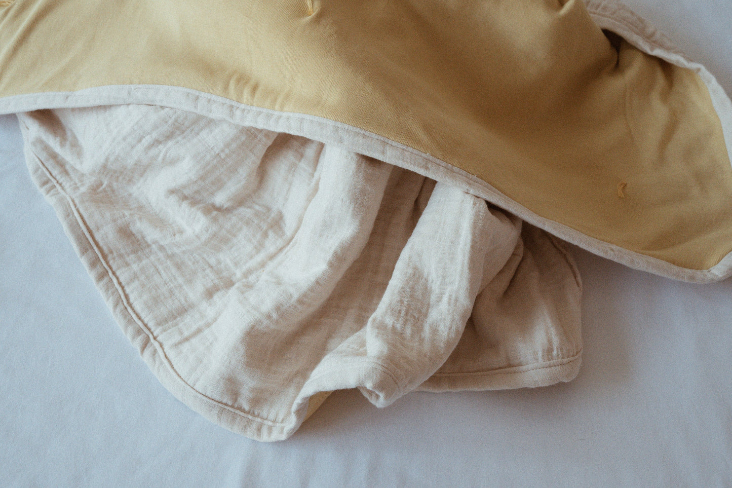 A close-up of two crumpled cabane childrenswear pre-order BABY BLANKETs in camomile, one beige and one off-white, lying on a white surface with soft natural light.