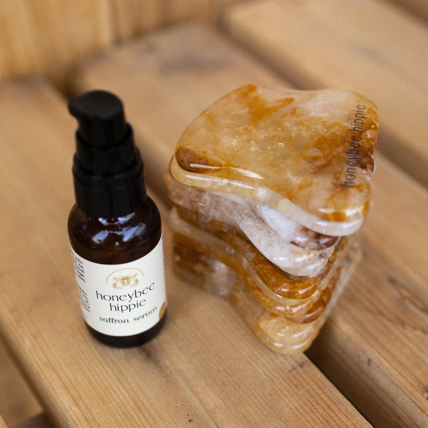 A small Honeybee Hippie Saffron Serum bottle—an antioxidant-rich blend for hyperpigmentation—stands next to orange and white stone gua sha tools on a wooden surface.
