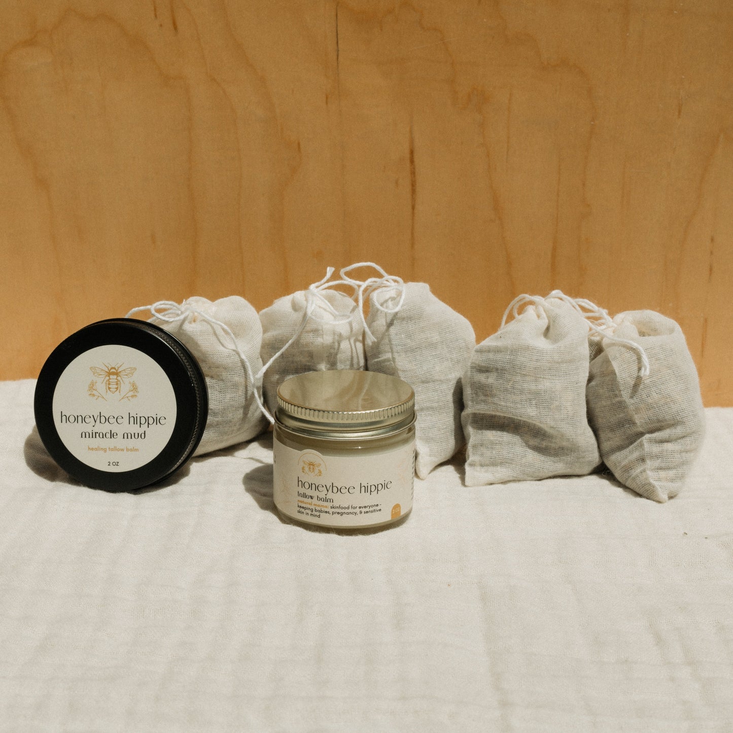 Two jars from the Honeybee Hippie "the natural mama bundle" sit against a wooden backdrop, flanked by small beige drawstring pouches, capturing a rustic and pure aesthetic.