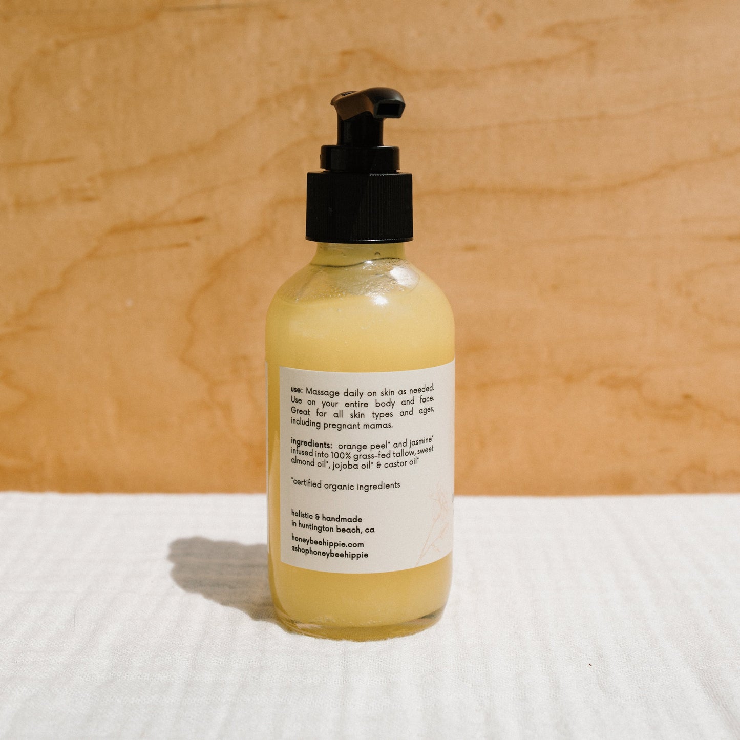 A glass pump bottle of Honeybee Hippie's "california sunshine - tallow oil," containing yellow lotion with organic orange peel, stands on a textured surface against a neutral background. The label lists uses and ingredients, featuring a non-comedogenic formula that nourishes without clogging pores.