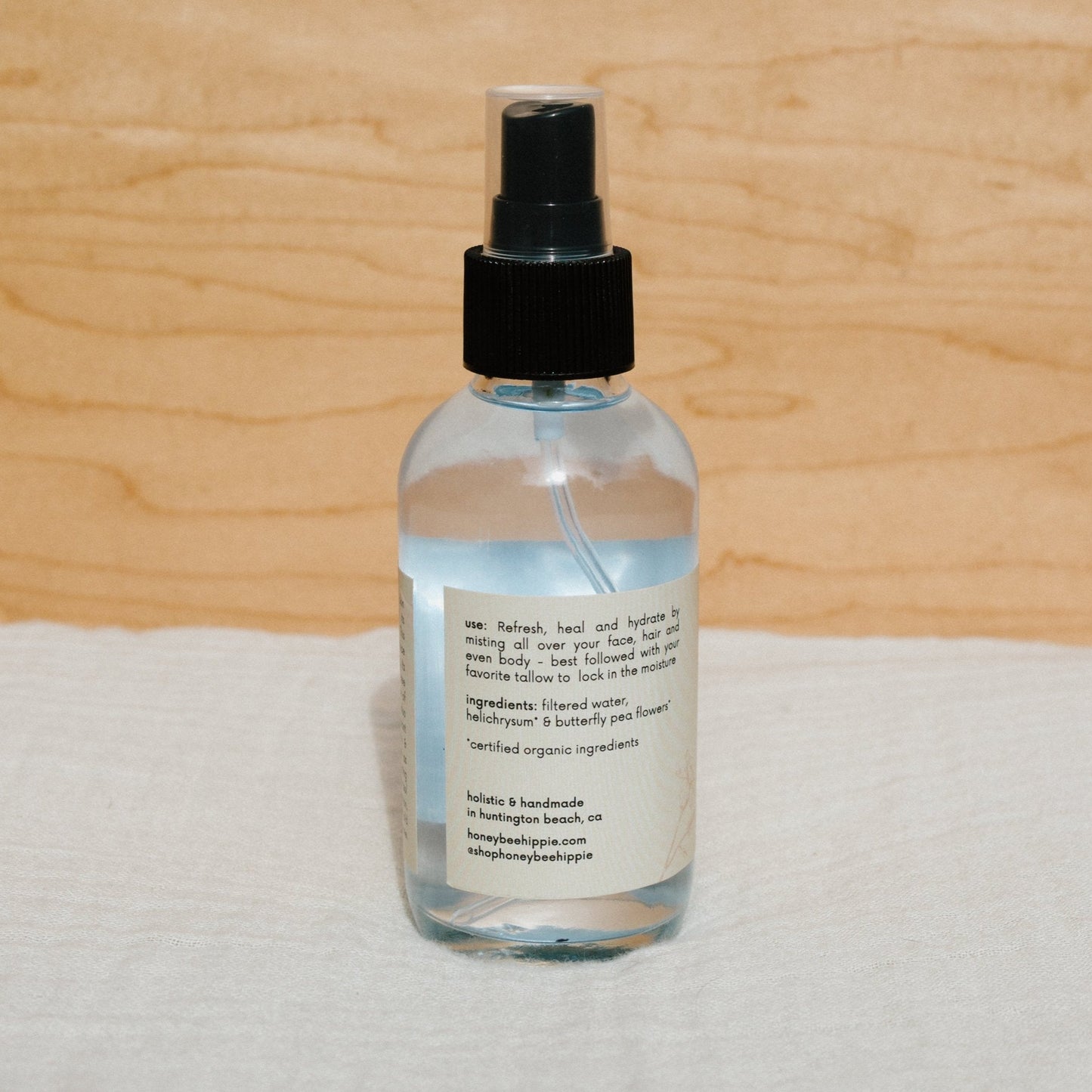 A Butterfly Hydrosol spray bottle from Honeybee Hippie features a black nozzle and label with usage instructions and ingredients like filtered water and helichrysm. The bottle rests elegantly on a soft, light-colored surface.