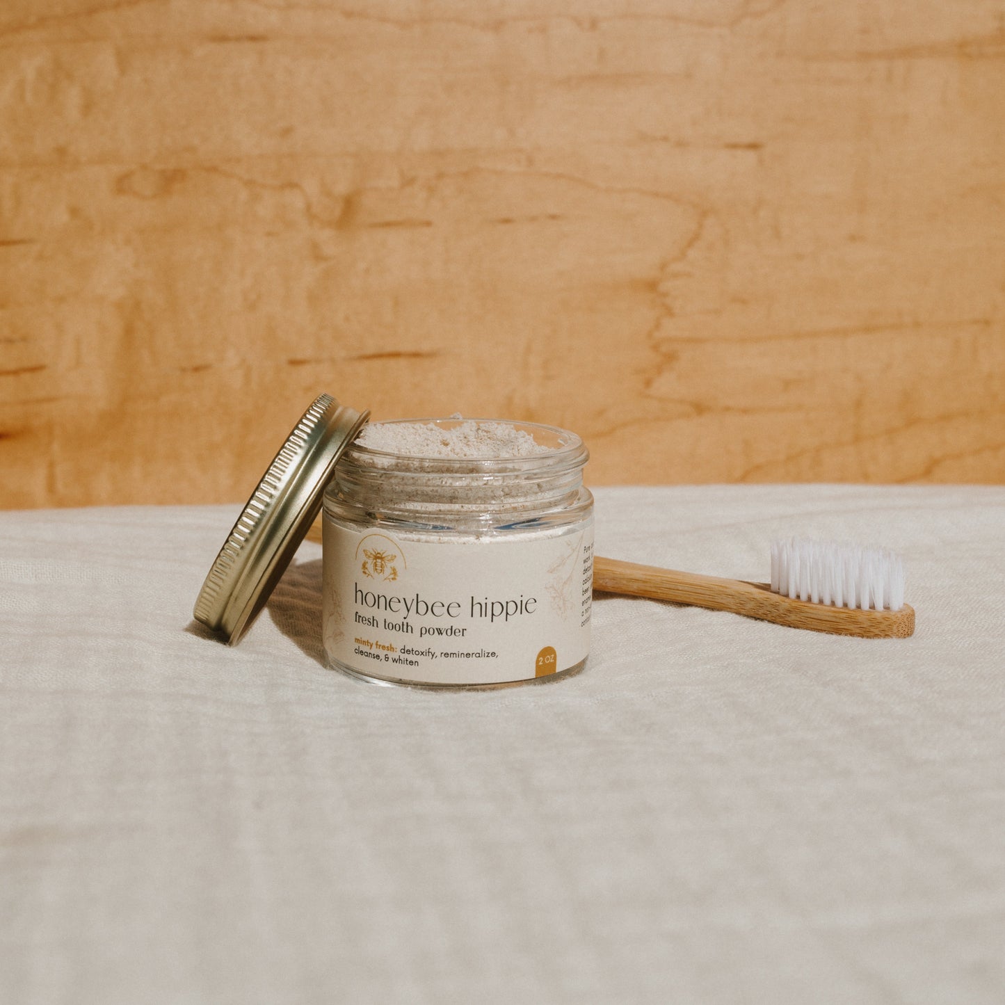 A "Honeybee Hippie fresh tooth powder" jar, known for remineralizing teeth, sits with its lid against it. Nearby, a wooden toothbrush with white bristles rests on pale fabric against a light wood background.