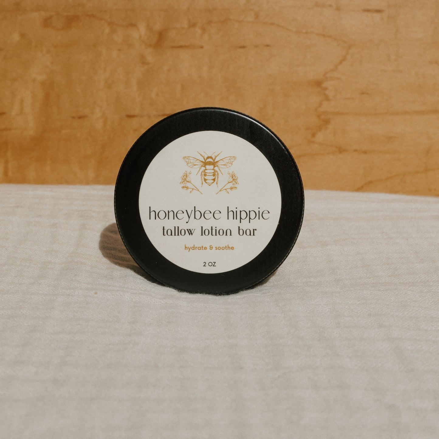 A black tin container featuring a bee illustration labeled "tallow lotion bar" by Honeybee Hippie rests on a wooden surface, boasting "hydrate & soothe with all-natural moisturizer, 2 oz.