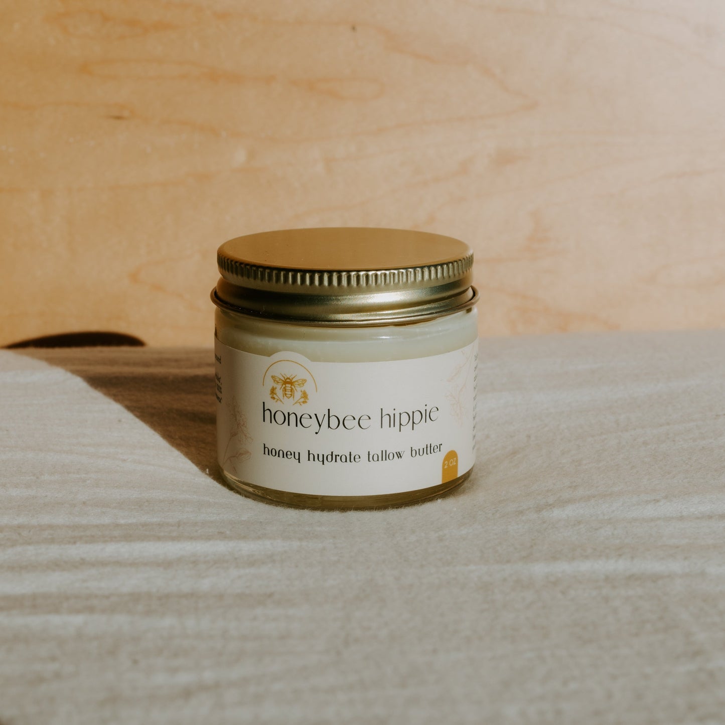 A jar of Honeybee Hippie's honey hydrate tallow butter rests on a light surface enriched with organic herbs. The label displays a bee illustration, and the metallic gold lid shines against the smooth wooden backdrop.