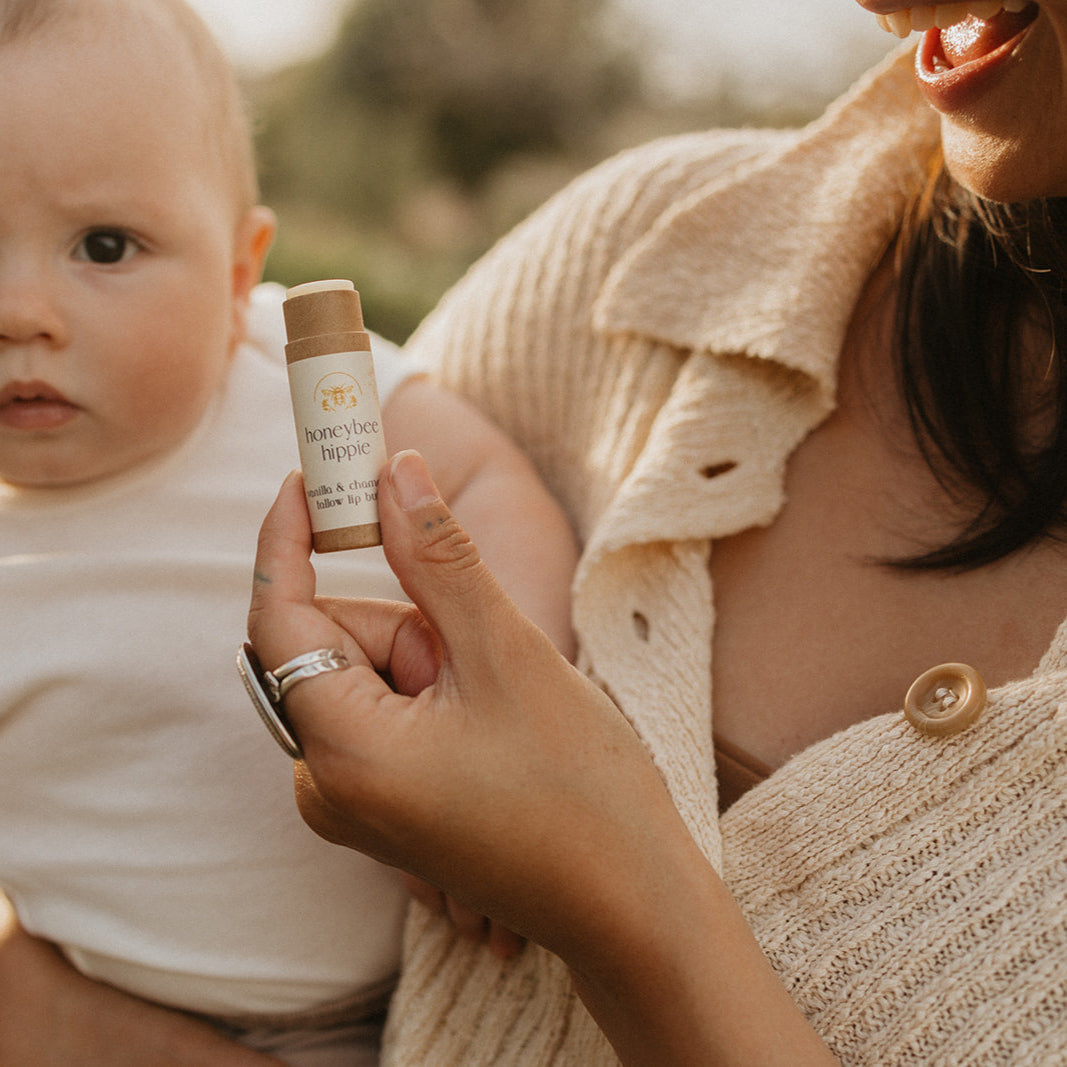 A person with a baby on their lap holds a tube labeled "vanilla & chamomile tallow lip butter" by Honeybee Hippie. Both wear light-colored clothing, set amidst soft lighting and greenery, highlighting the product.