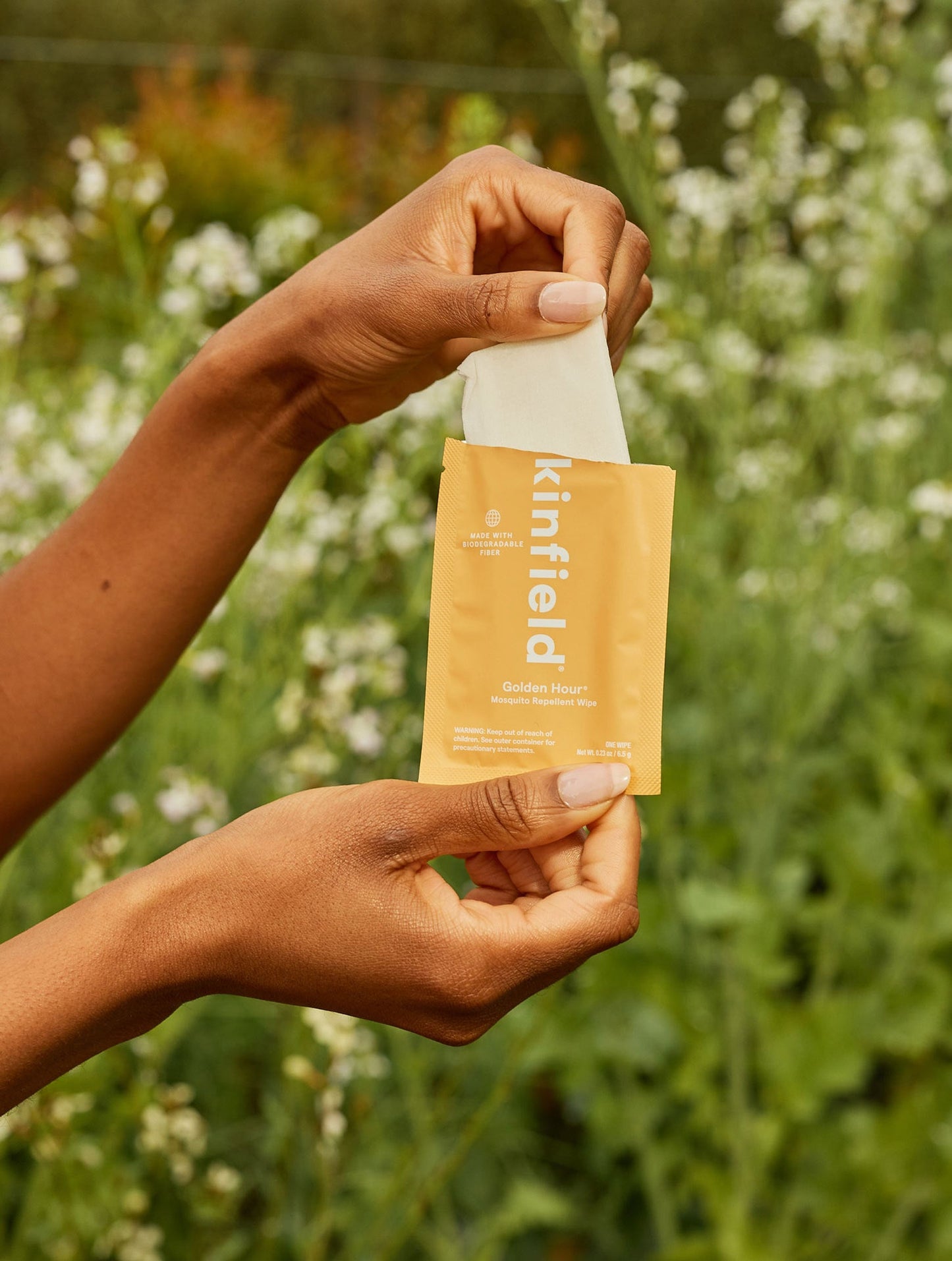 Two hands opening a yellow Kinfield Golden Hour™ Wipes packet in front of a green, blurred background with white wildflowers. The travel-friendly mosquito repellent packet likely contains Indonesian citronella wipes. The person's skin is medium-brown, and the setting appears to be outdoors.