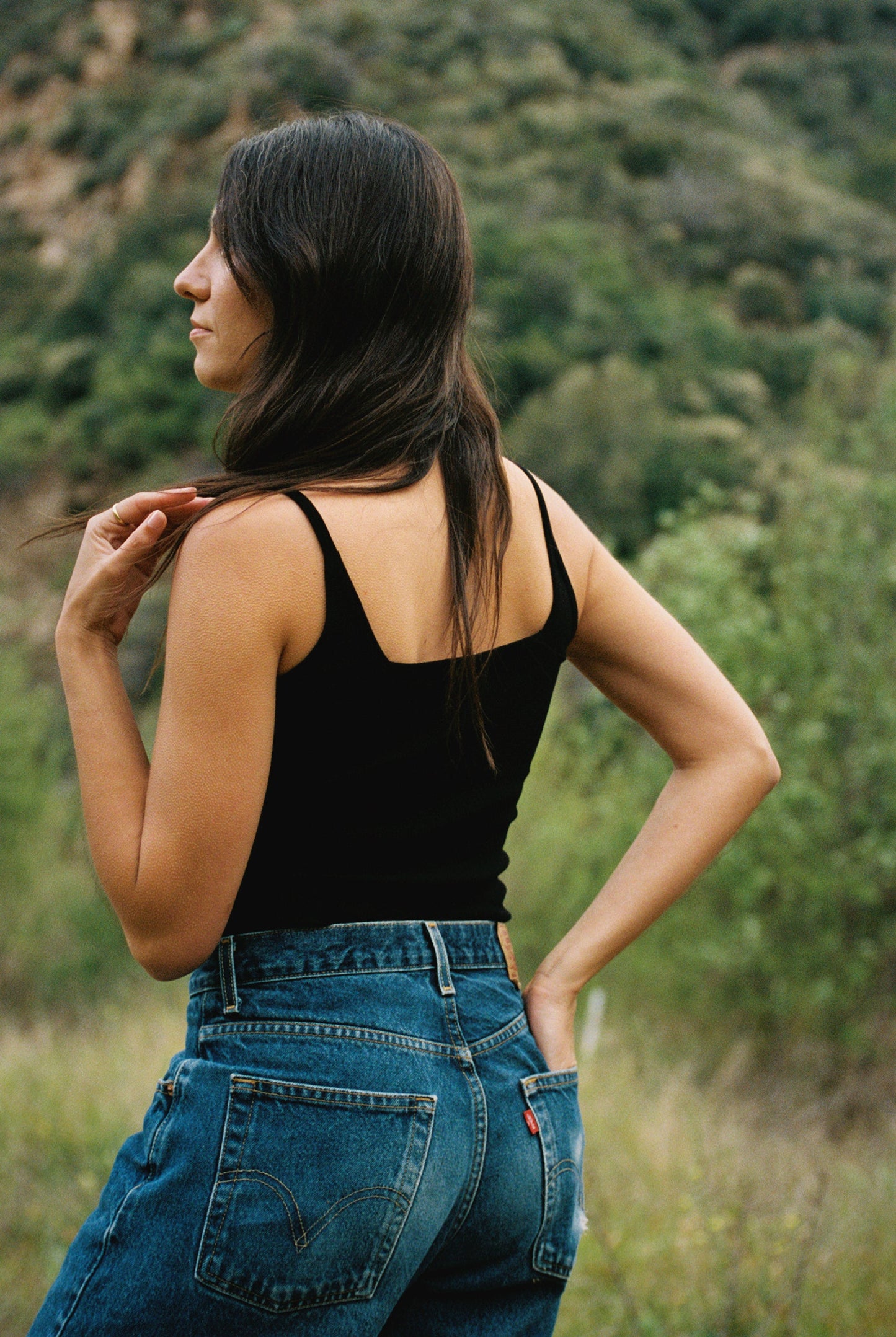 In Los Angeles, a woman with long dark hair stands outdoors, gazing to the left. She's wearing LA Relaxed's Organic Square Cami and blue jeans. Her right hand rests on her hip as her left touches her hair, with green foliage and hills in the background.