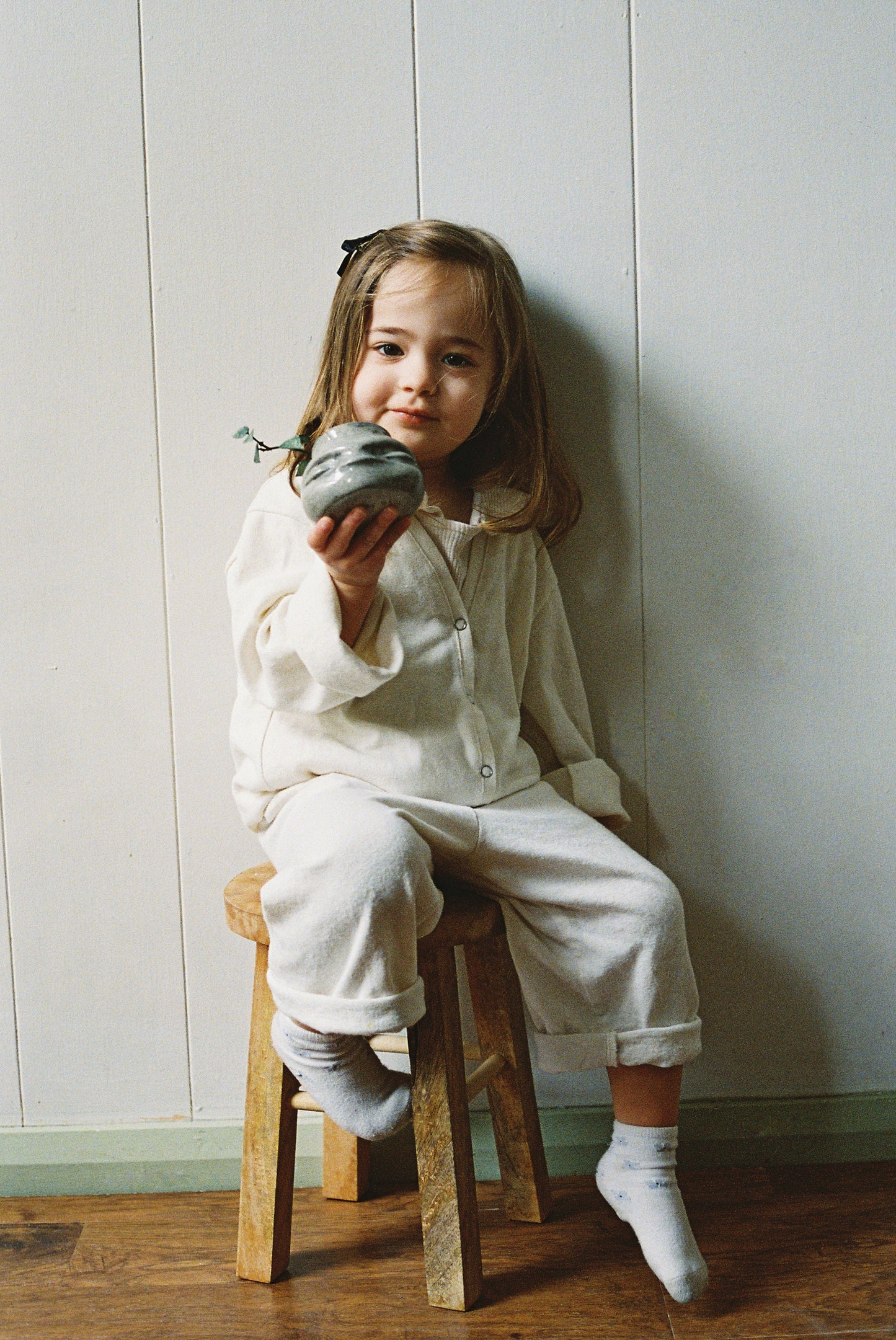 A young girl with shoulder-length hair and socks sits on a wooden stool against a white wall, smiling gently at the camera while wearing the TAKE CARE TOP hemp by cabane childrenswear and holding a small potted plant.