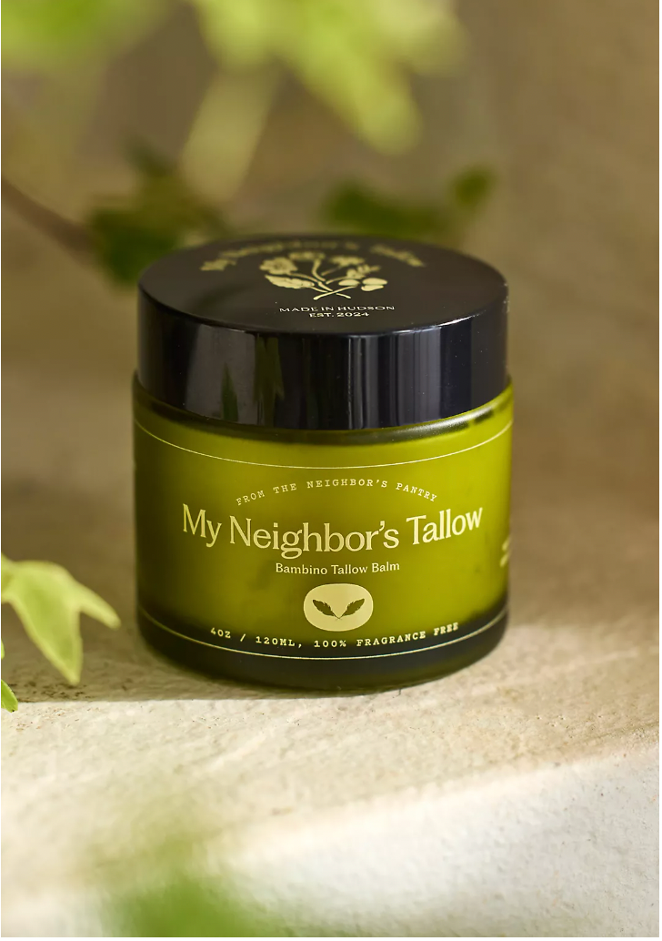 A green jar of My Neighbor’s Tallow Bambino Balm - Fragrance Free Tallow (4oz/120mL) sits on a light stone surface with green leaves. Made from grass-fed tallow, it's 100% fragrance-free and ideal for sensitive skin.