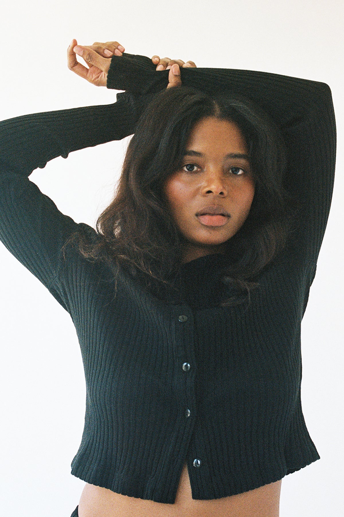 A woman with long, wavy hair wears the LA RELAXED Pointelle Cardigan, a black ribbed button-up with pearlescent buttons. She poses against a plain background, arms raised and crossed above her head, gazing at the camera with a relaxed look.