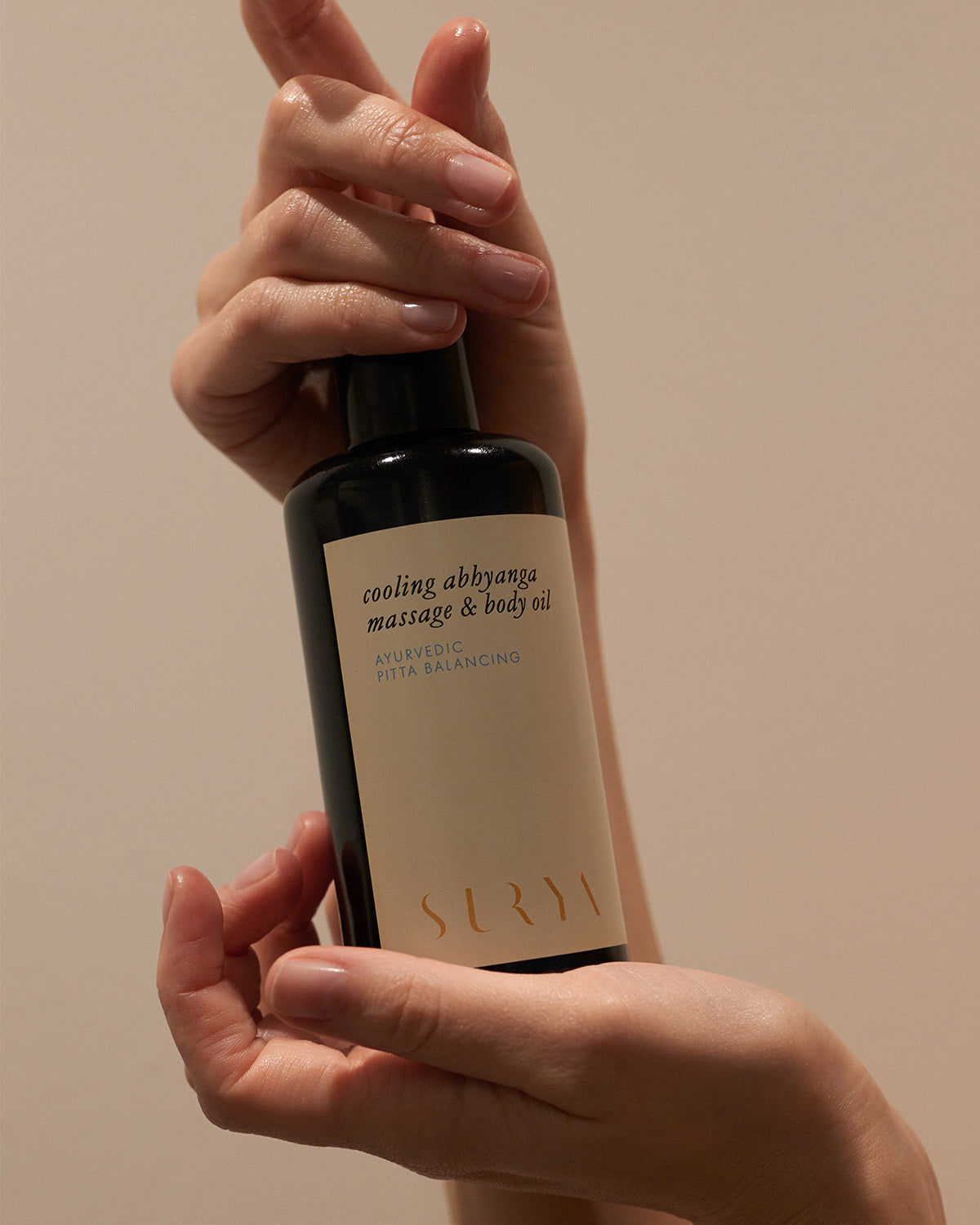 Hands holding a dark bottle with a beige label that reads "cooling body oil, herbally rich blend for ayurvedic pitta balancing." The brand name "Surya" is printed at the bottom of the label.