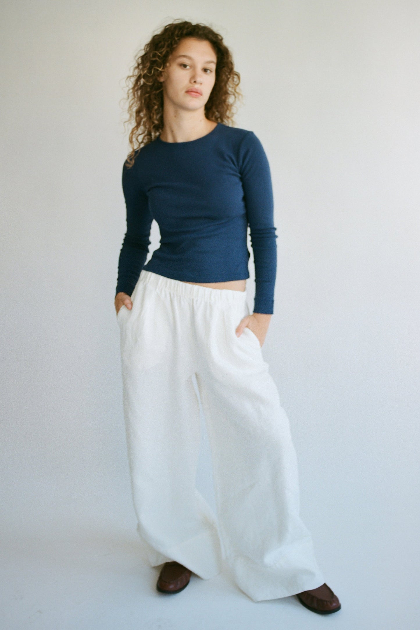 A person with curly hair stands against a plain background, wearing a LA RELAXED Bebe Organic Long Sleeve navy top and loose pants. Their hands are in their pockets as they look at the camera.