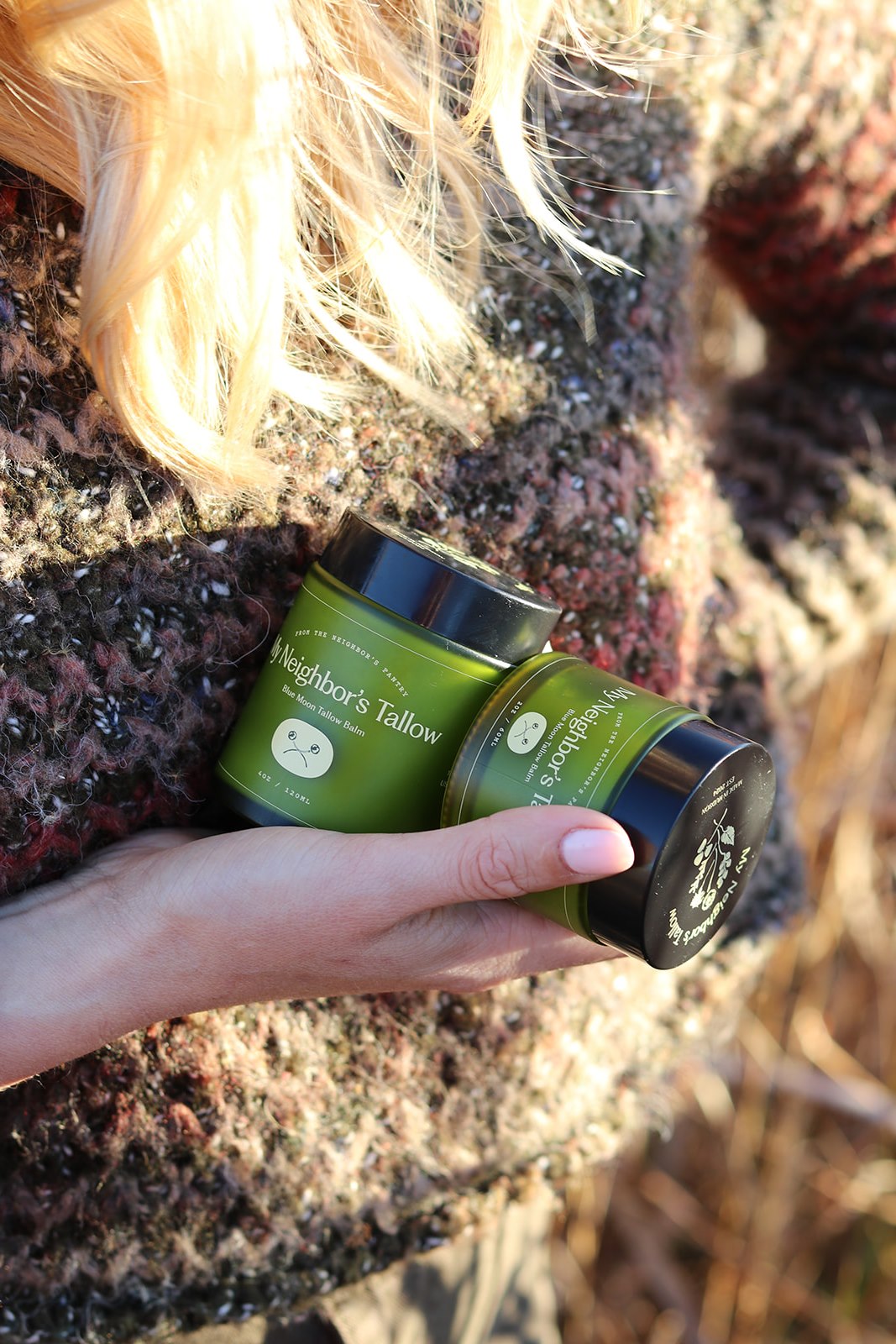 A person with long blonde hair, wearing a textured sweater, holds two green jars labeled "Blue Moon Face Balm" by My Neighbor’s Tallow—a natural face moisturizer—outdoors in natural sunlight.