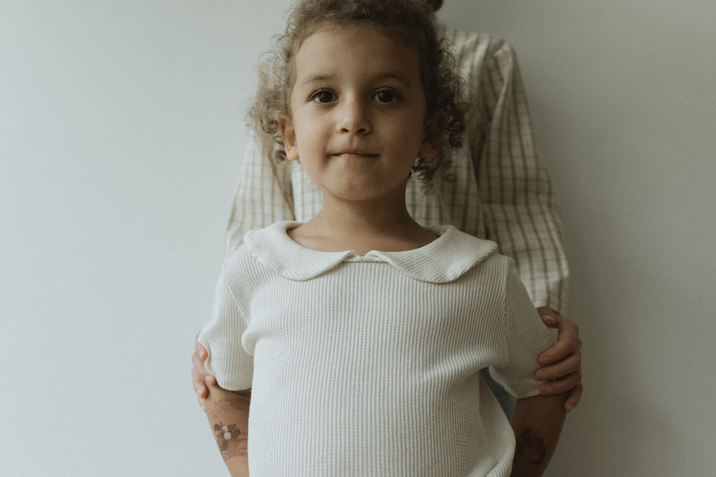 A young child with curly hair poses in a SCHOOLHOUSE TEE waffle by cabane childrenswear, embodying classic looks. They stand before an adult in a plaid shirt with hands resting gently on the child's shoulders, all set against a plain wall that highlights their connection and timeless appeal.