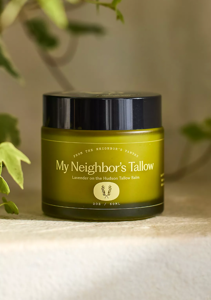 A green jar of My Neighbor’s Tallow "Lavender on the Hudson" Tallow Balm sits on a surface, leafy plants blurred in the background, hinting at its blend of nourishing grass-fed tallow and jojoba oil.