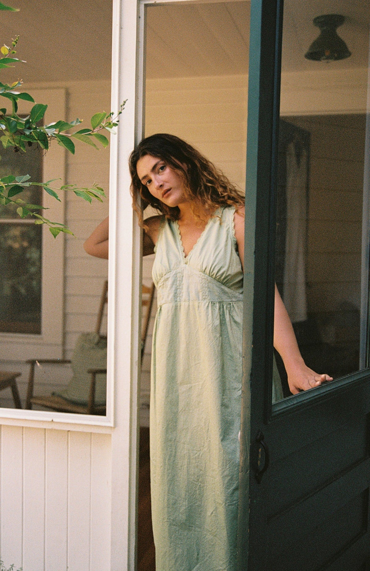 Mint - A woman with wavy hair stands in a doorway, one hand to her head, wearing the LA RELAXED Makinna Poplin Dress in light green organic cotton. Sunlight filters through nearby greenery as she gazes out with a relaxed expression.