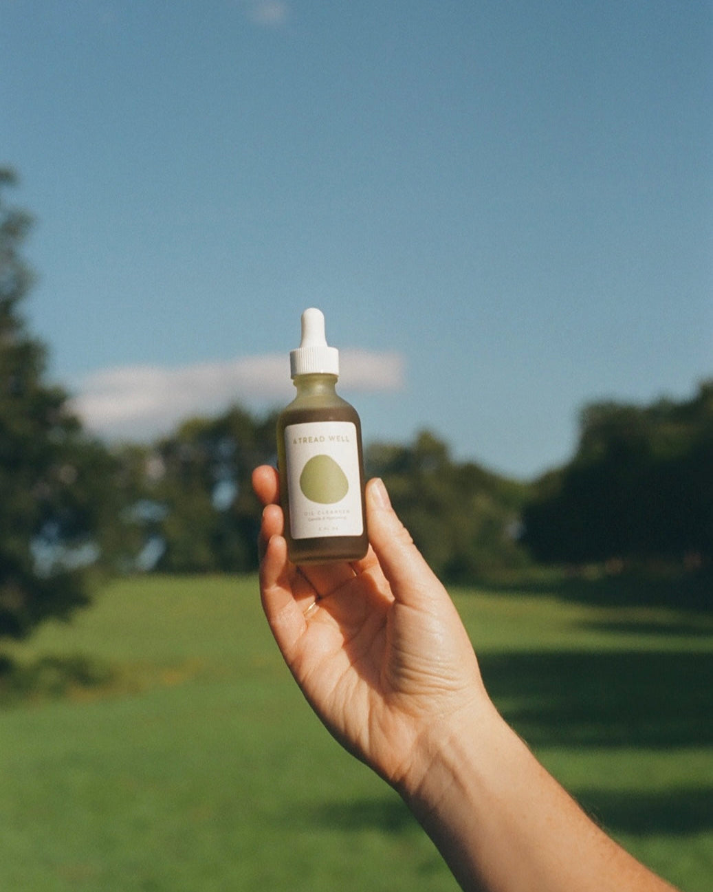 A hand holds up a bottle of & Tread Well Oil Cleanser with a dropper lid against a blurred backdrop of green grass, trees, and blue sky.