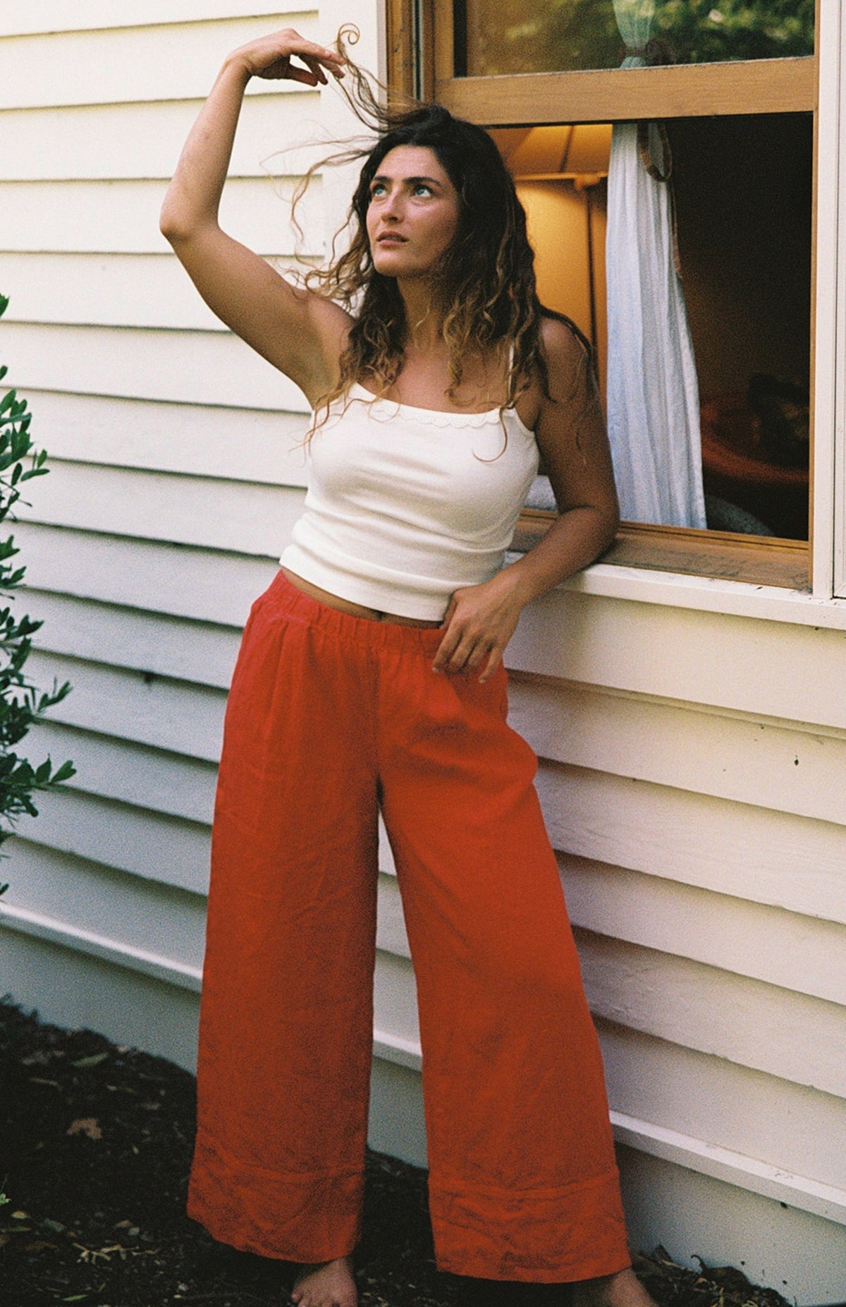 Cream -
A woman wears the LA RELAXED Frill Camisole and loose red pants, standing outside by a white wooden building, leaning against a window and looking up while twirling her hair.