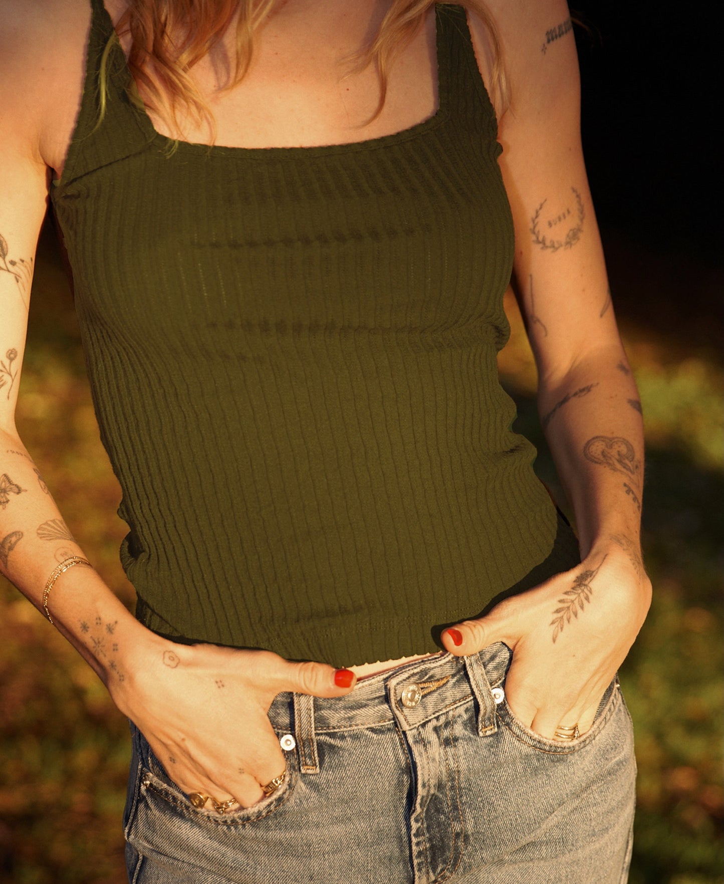 Sage Wearing the LA Relaxed Pointelle Square Cami in green and blue jeans, a person with tattooed arms stands outdoors with hands in pockets as sunlight highlights their skin and clothing.
