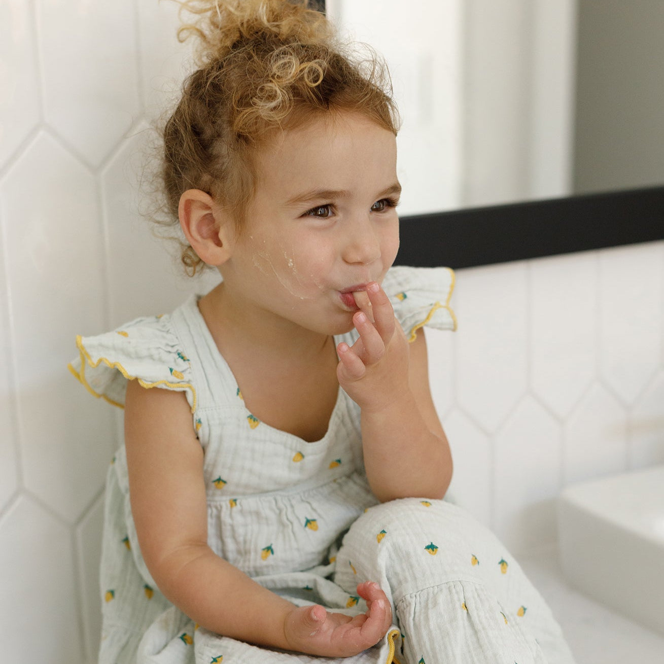 A young child with curly hair in a light dress sits by a sink, smiling and licking food from her fingers—a wholesome scene as pure as Wild Mamas’ Wildly Nourishing Bison Tallow Balm - Raw Honey for gentle skincare. White tiles and a mirror behind.