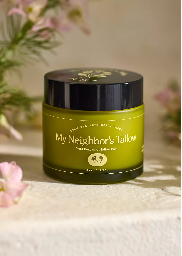A green 2 oz (60 mL) jar of My Neighbor’s Tallow Wild Bergamot – Tallow Balm rests on a light surface, with soft pink flowers and greenery blurred in the background. The balm is made from grass-fed tallow and wild bergamot.