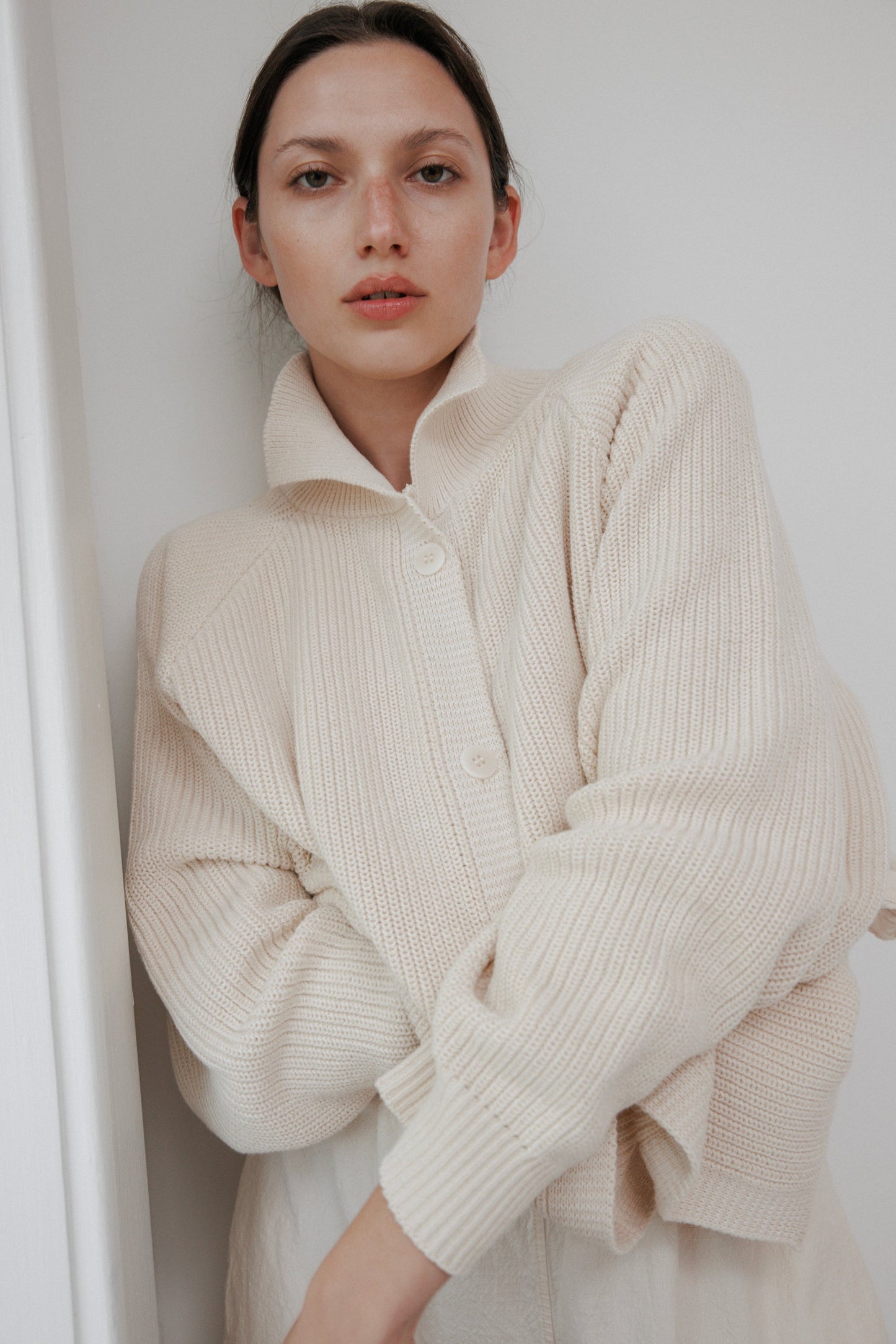 A woman stands against a white wall, arms crossed, wearing the Wol Hide Collar Cardigan: Natural. Her neutral expression and dark hair pulled back blend with the lightweight knit's minimalistic style, creating a serene look.
