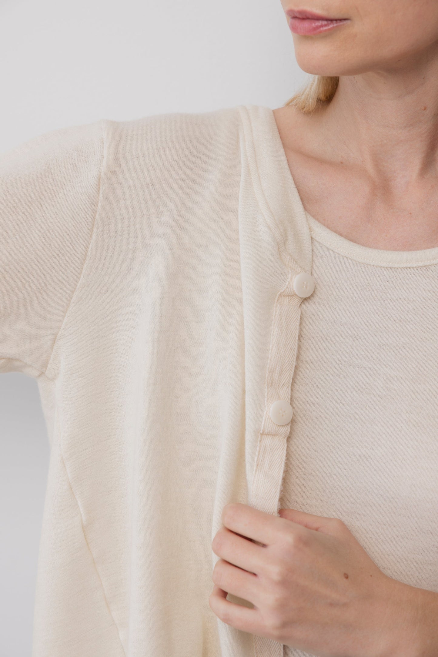 A woman wearing the Wol Hide Wool Liner in Natural fastens its large Corozo buttons, with only her lower face, neck, and upper body visible against a plain white background.