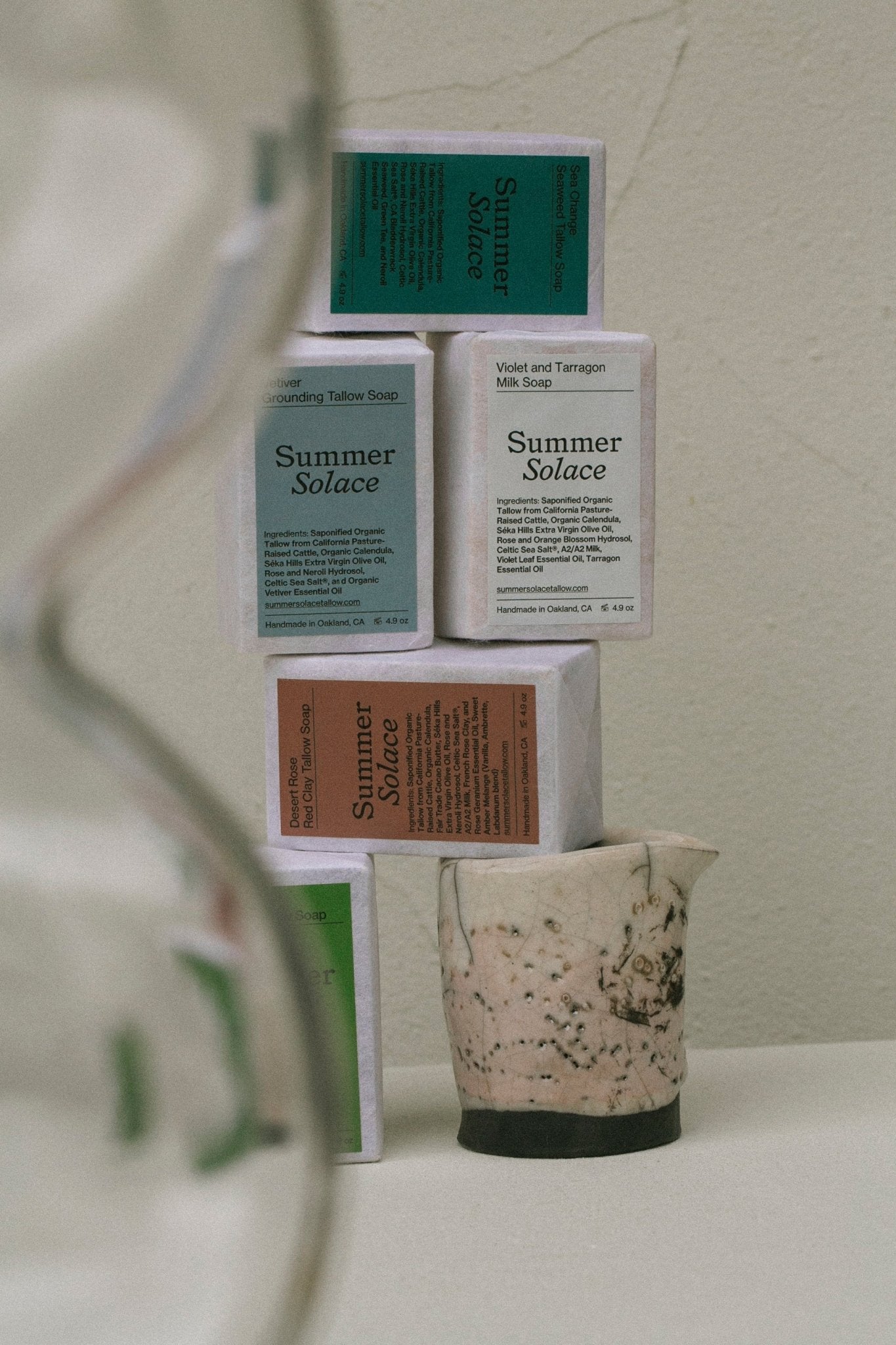 An arrangement of five Desert Rose Red Clay Bar Soaps in green, white, and brown packaging from Summer Solace Tallow is stacked on top of a marbled ceramic cup. An out-of-focus hourglass is partially visible in the foreground to the left. The background features a neutral-toned wall. The Regenerative Tallow™ and French rose clay add a luxurious touch.