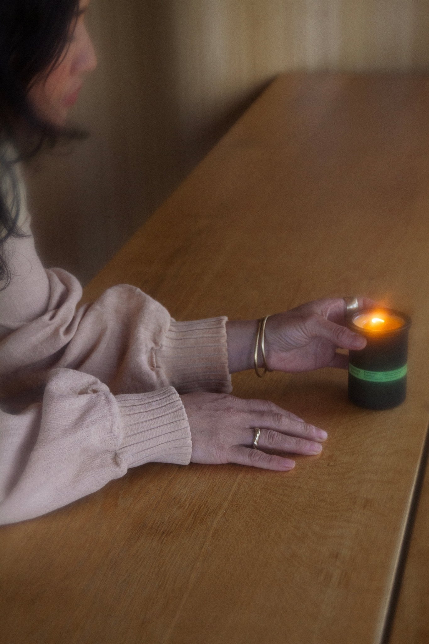 A person with long hair and a peach sweater holds an Evergreen Essence (Cedar and Oakmoss) Candle - Regenerative Tallow® by Summer Solace Tallow on a wooden table. The lit candle casts a warm glow, filling the softly focused scene with a calm, woodsy fragrance that enhances the serene atmosphere.