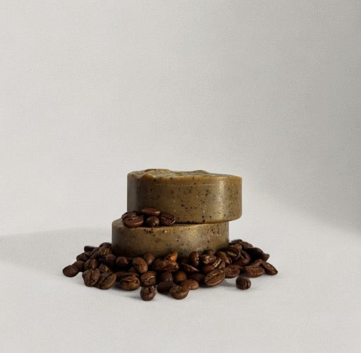 Two round bars of Wild Mamas' Wild Bison Tallow Deep Exfoliating Soap Scrub—Shilajit + Decaffeinated Coffee Bean—are stacked with coffee beans scattered around, highlighting the regenerative ingredients, against a plain white background.