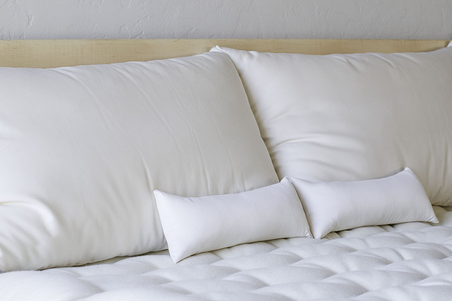 A neatly made bed features a tufted white mattress and smooth white pillows, including Shepherd's Dream Neck Pillow. The light wood headboard complements the two large pillows and two smaller ones, all in a uniform white color, creating a clean and minimalist look.