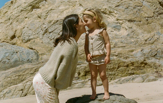 Founder Nikki touching noses with her daughter on the beach with rocks and blue sky behind