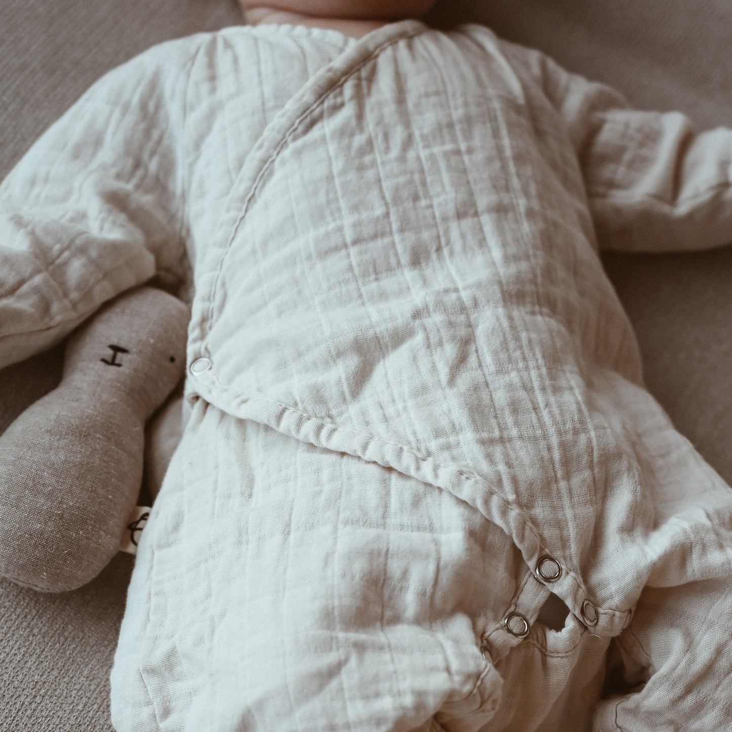 A baby is comfortably dressed in the Après Organic Wrap Undyed by cabane childrenswear, made from breathable fabric, and lies on a neutral-toned surface with a minimalistic stuffed animal toy nestled beside them.