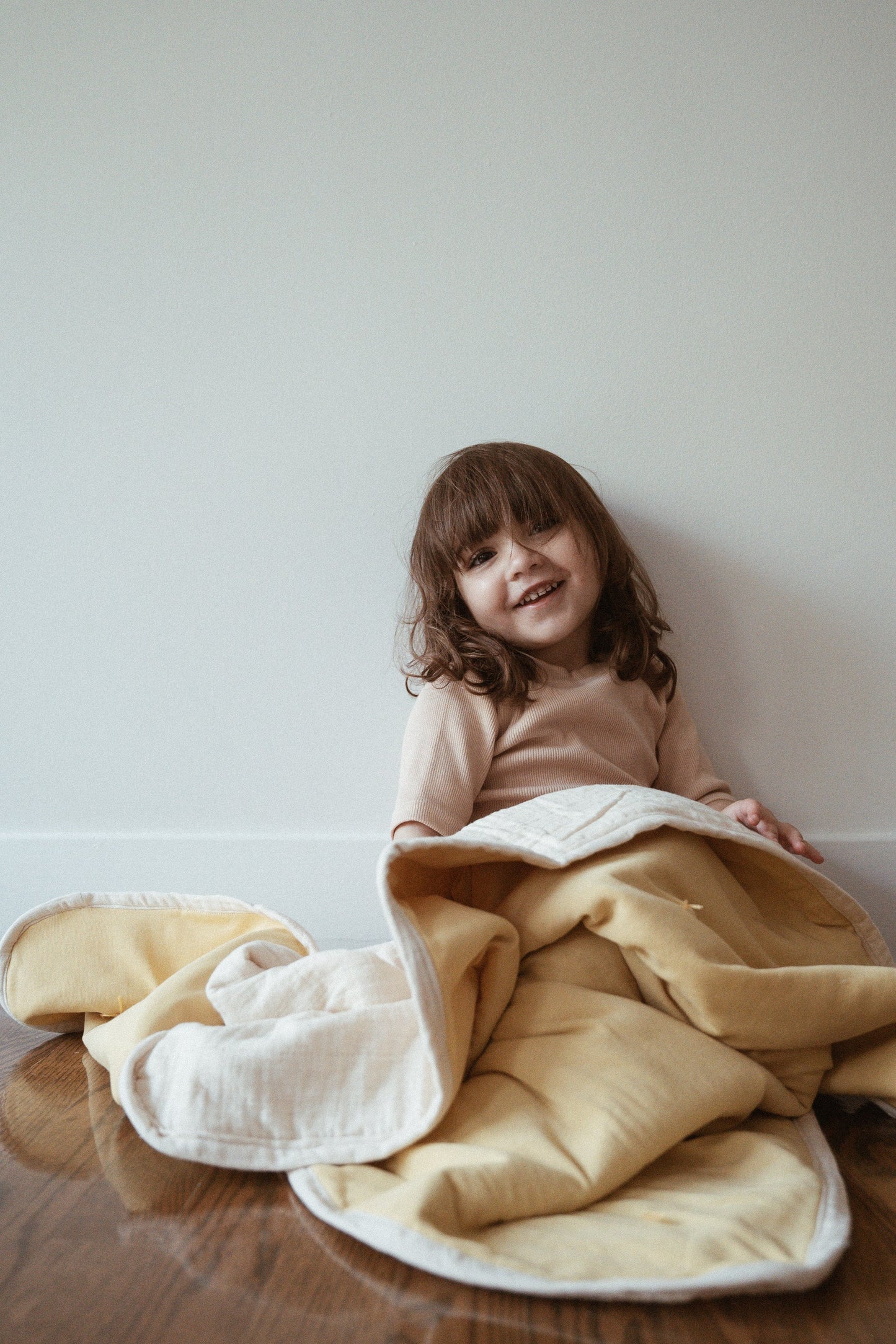 A young child with brown hair sits smiling on a wooden floor, wrapped in a cozy yellow and white camomile pre-order BABY BLANKET by cabane childrenswear, against a plain white wall.