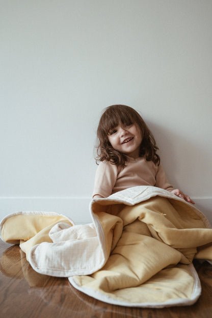 A young child with brown hair sits smiling on a wooden floor, wrapped in a cozy yellow and white camomile pre-order BABY BLANKET by cabane childrenswear, against a plain white wall.