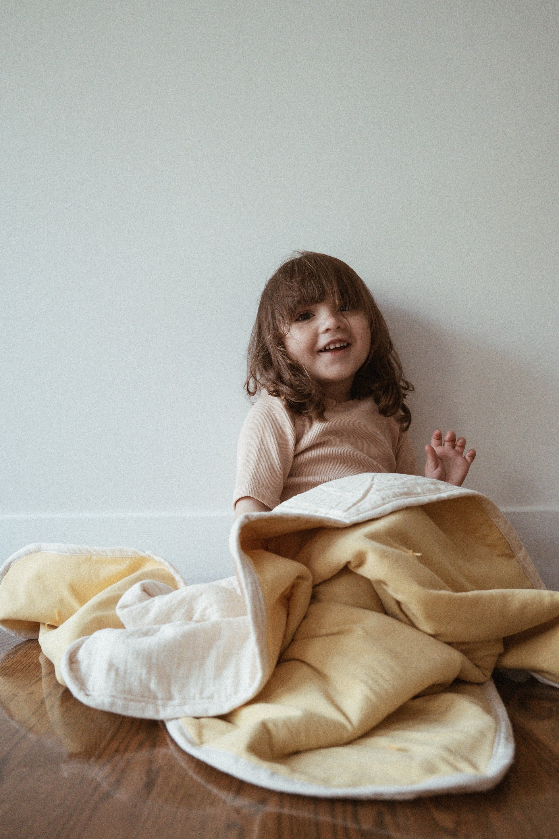 A young child with long brown hair smiles on a wooden floor, partially wrapped in the cabane childrenswear pre-order BABY BLANKET, camomile. The hand-quilted yellow and white blanket stands out against the plain white wall behind.