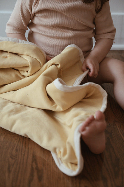 A young child sits on a wooden floor, holding the cabane childrenswear pre-order BABY BLANKET in camomile, its soft yellow hand-quilted fabric with white edges complementing their light ribbed outfit. The baby's face is not visible.
