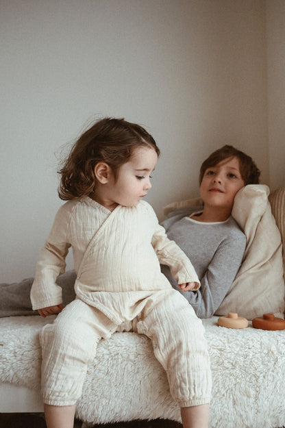 Two children sit on a bed in a cozy room. The younger child, wrapped in an Après Organic Wrap Undyed from cabane childrenswear, looks down, while the older child, wearing a gray top made from breathable fabric, reclines with a pillow behind them. Wooden toys are scattered across the bed.