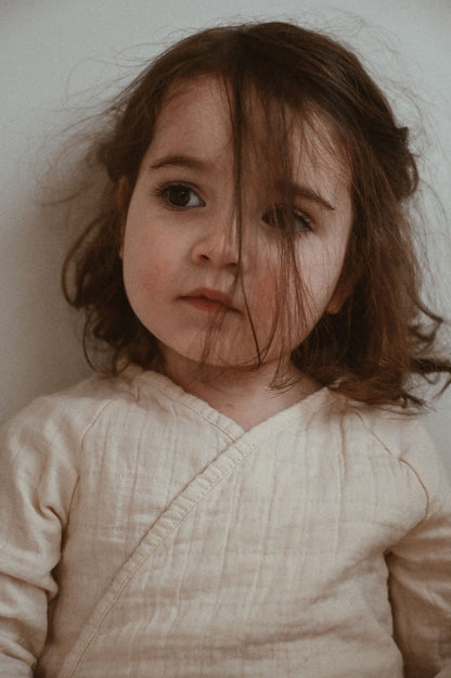 A young child with long brown hair, wearing the light and breathable Après Organic Wrap Undyed by cabane childrenswear.