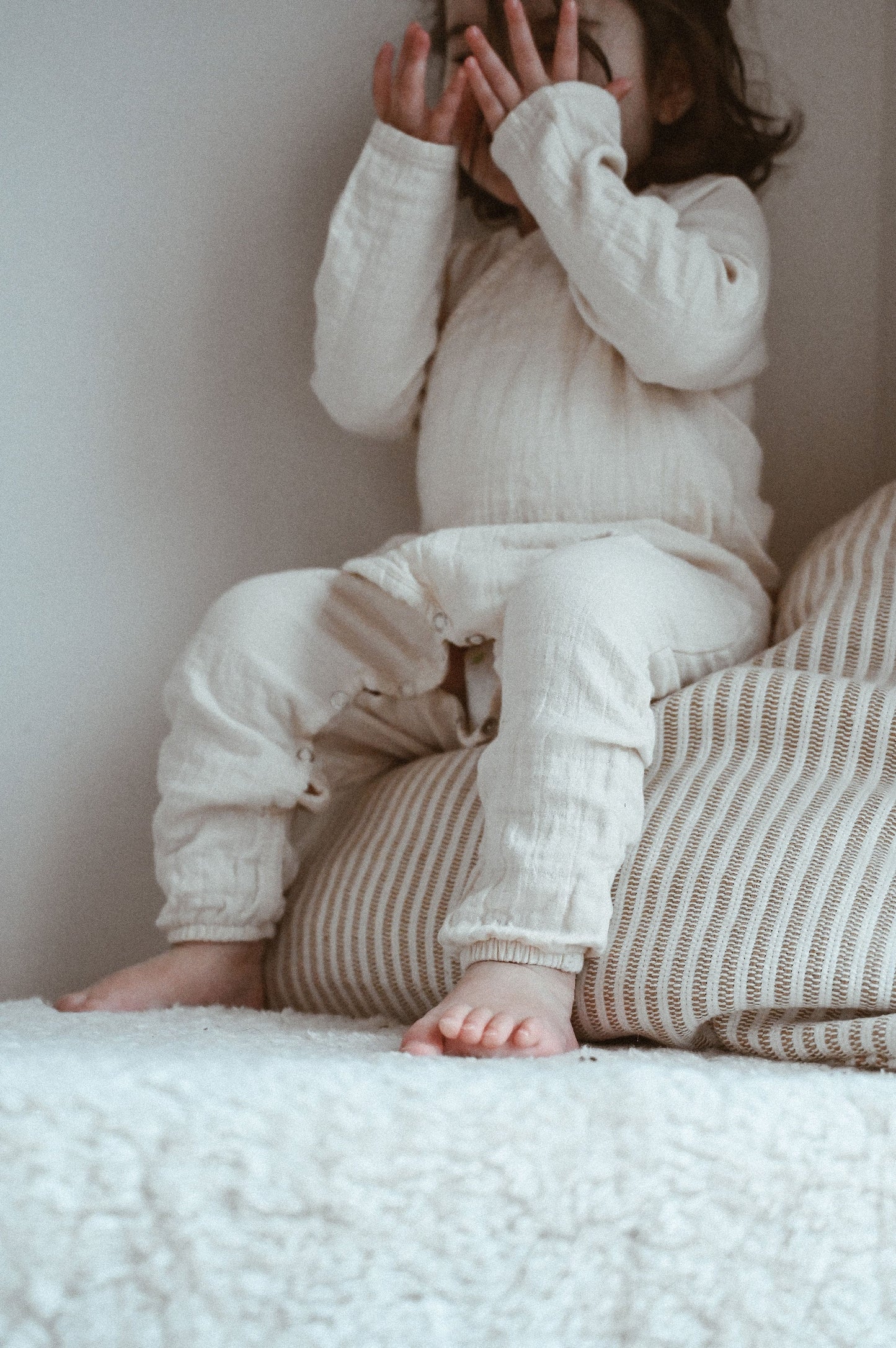 A child dressed in the Après Organic Wrap Undyed from cabane childrenswear is seated on a striped cushion against a soft backdrop, playfully hiding their face with their hands. 
