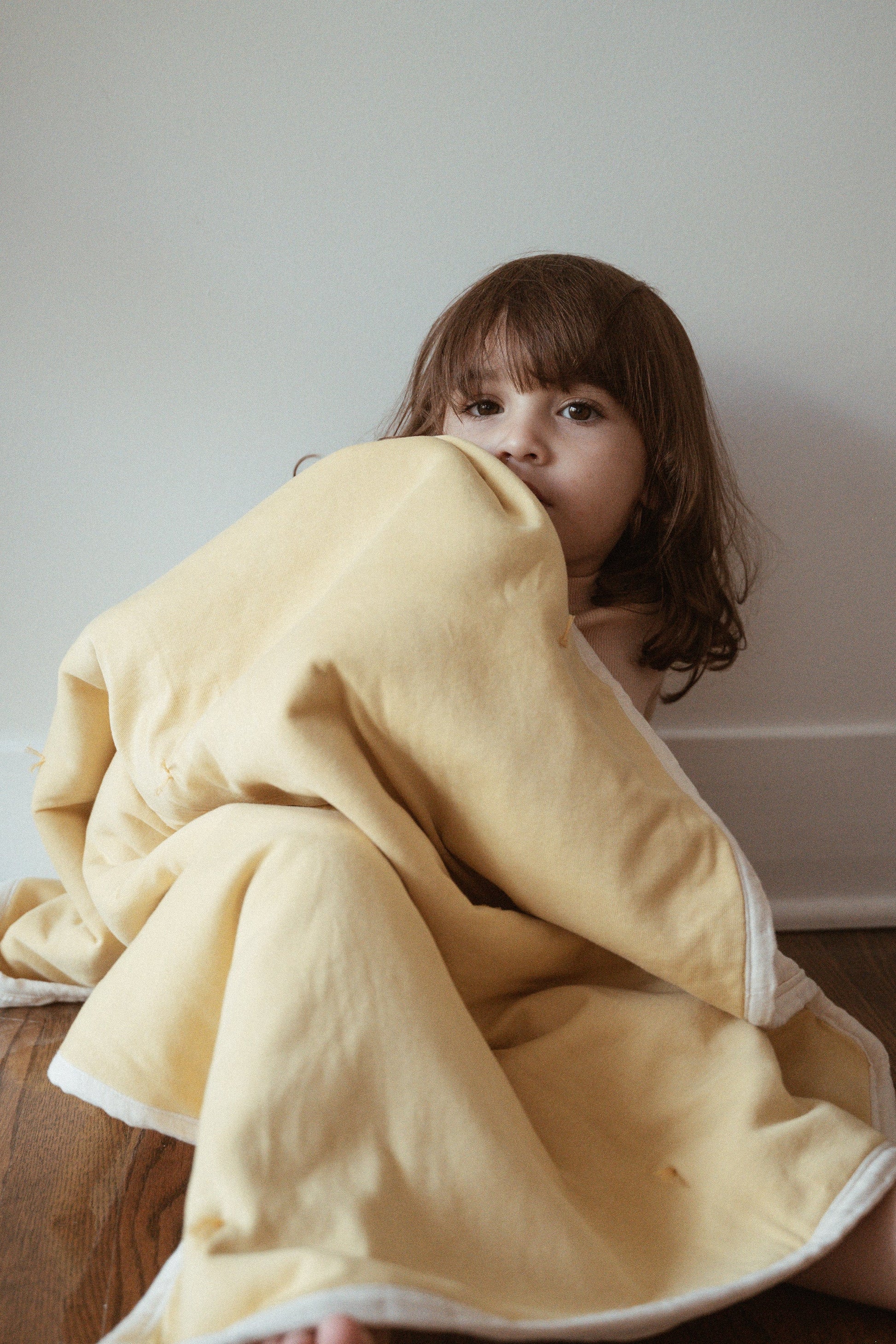A young child with brown hair sits on a wooden floor, partially wrapped in the cabane childrenswear pre-order BABY BLANKET in camomile, looking thoughtfully at the camera against a plain white wall.