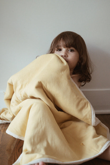 A young child with brown hair sits on a wooden floor, partially wrapped in the cabane childrenswear pre-order BABY BLANKET in camomile, looking thoughtfully at the camera against a plain white wall.