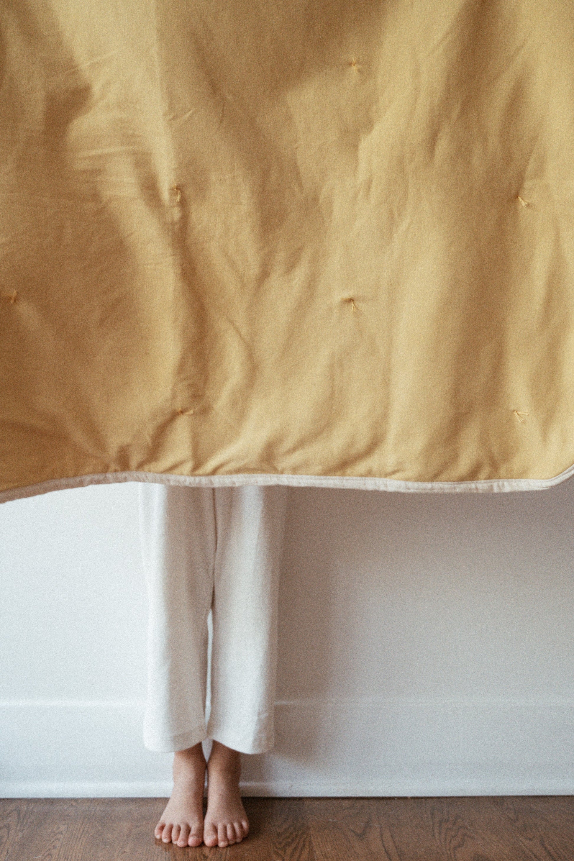 A person stands barefoot on a wooden floor, feet and lower legs visible below the hanging pre-order BABY BLANKET, camomile by cabane childrenswear. They are wearing loose white pants.
