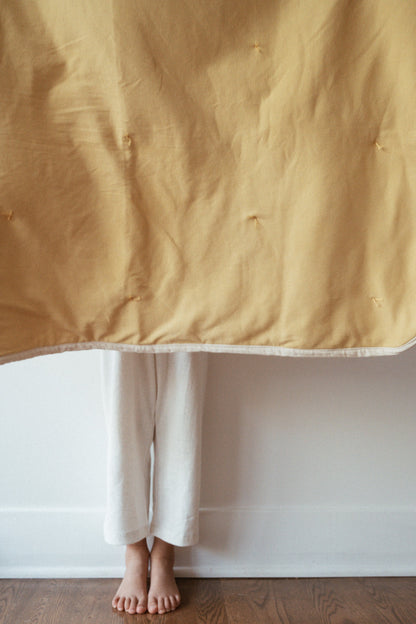 A person stands barefoot on a wooden floor, feet and lower legs visible below the hanging pre-order BABY BLANKET, camomile by cabane childrenswear. They are wearing loose white pants.