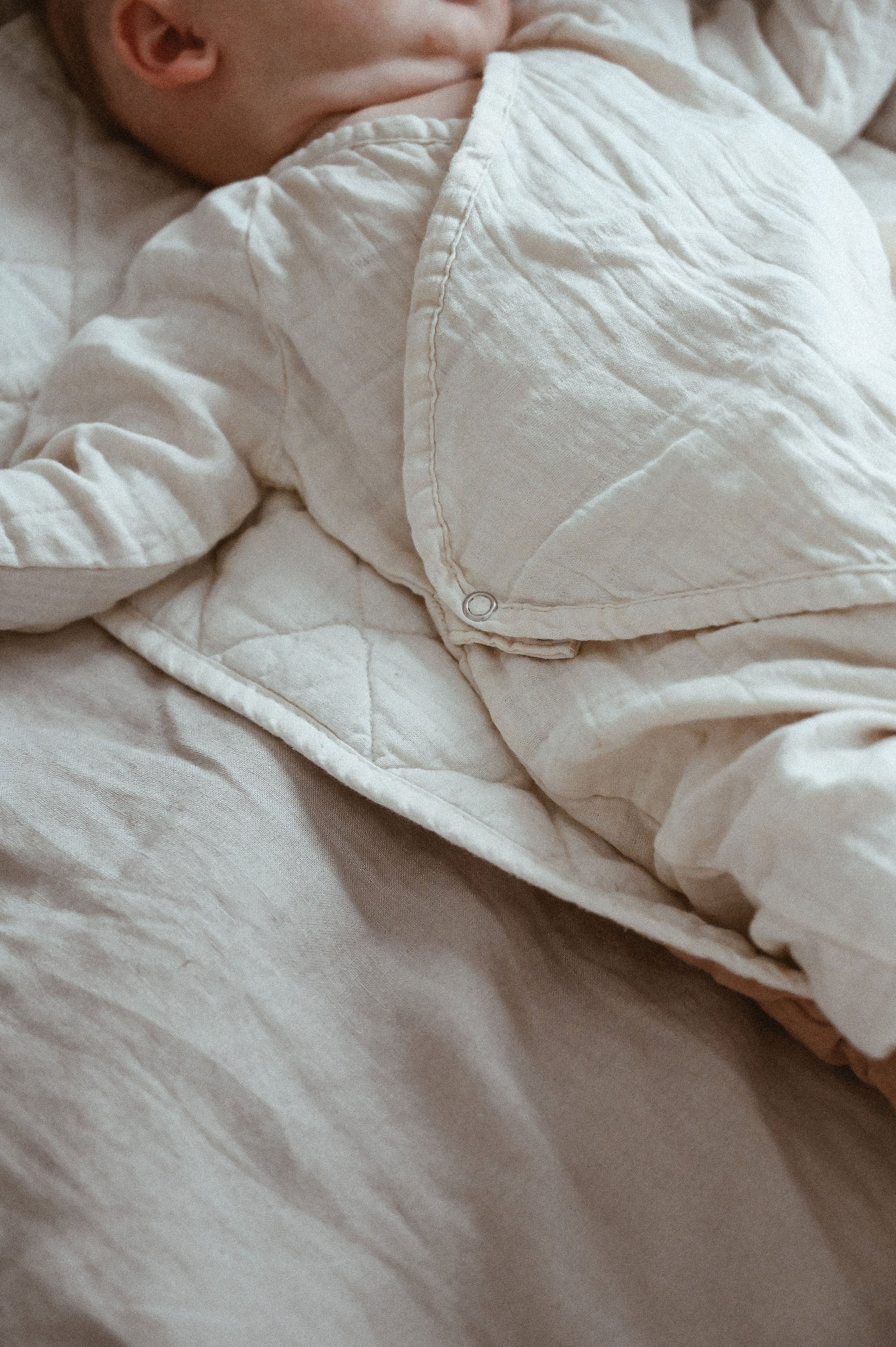 A baby is resting on a quilted blanket, wearing the Après Organic Wrap Undyed from cabane childrenswear. The outfit is crafted from breathable fabric, and only the upper body, including the baby's arm and the cozy garment with padded snaps, is visible.