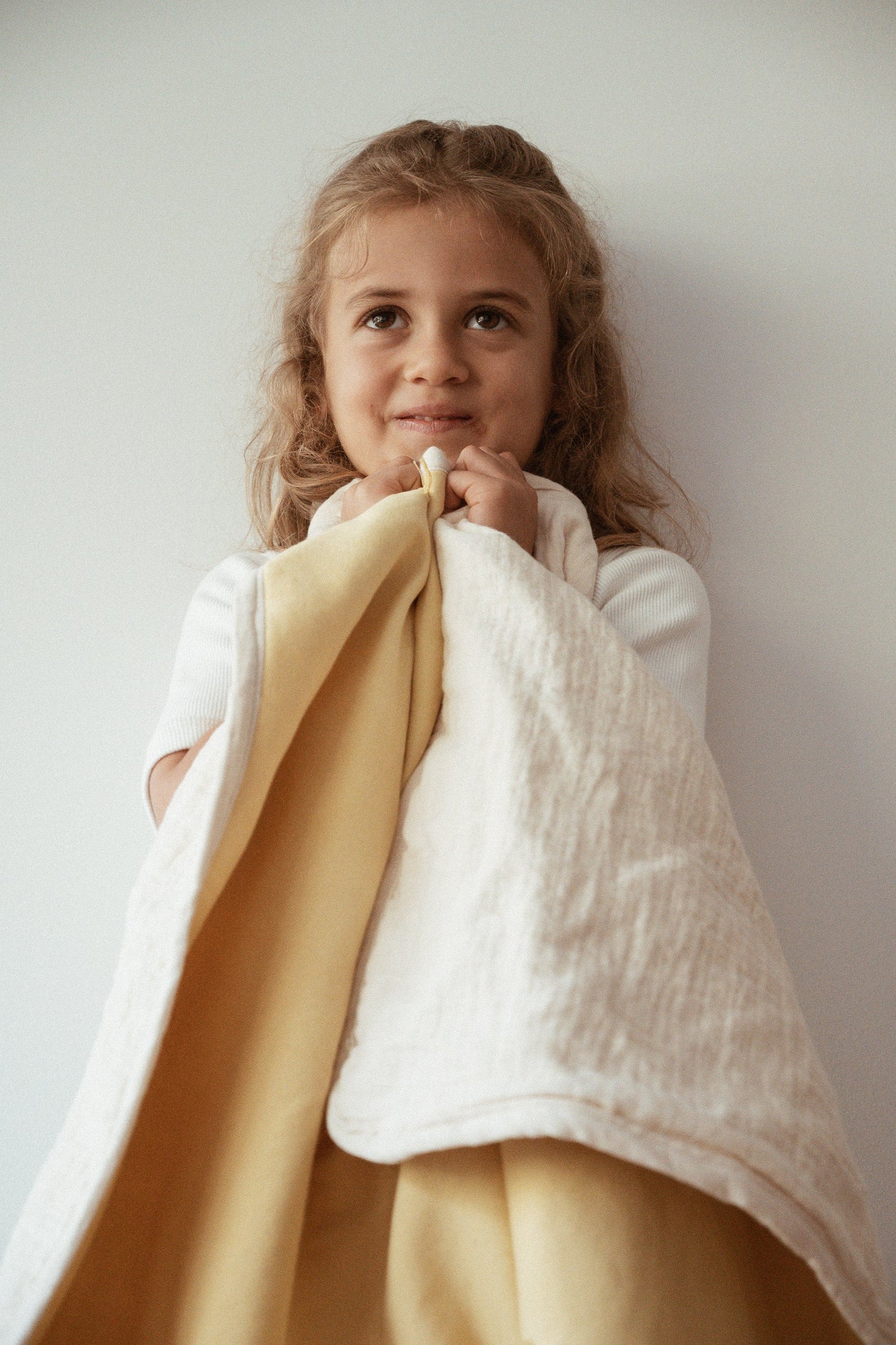 A young child with long, light brown hair smiles gently, holding the cabane childrenswear pre-order BABY BLANKET “camomile” with delicate marigold flowers against a plain white background.