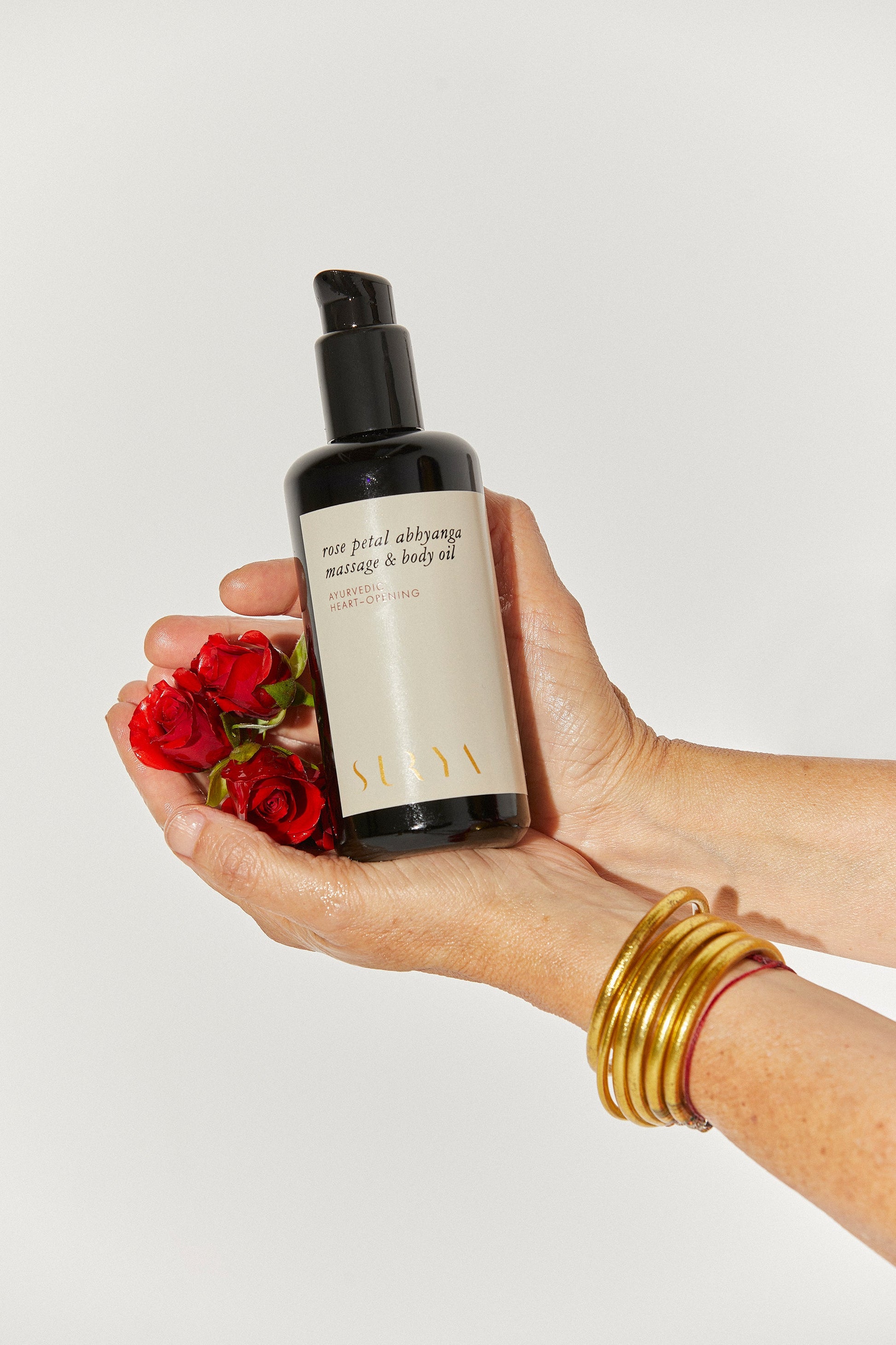 A hand adorned with gold bangles holds a black bottle of Surya's Rose Petal Body Oil, positioned beside a small bunch of red roses. The background is plain white, highlighting the bottle and flowers, emphasizing the organic ingredients used in this luxurious oil.