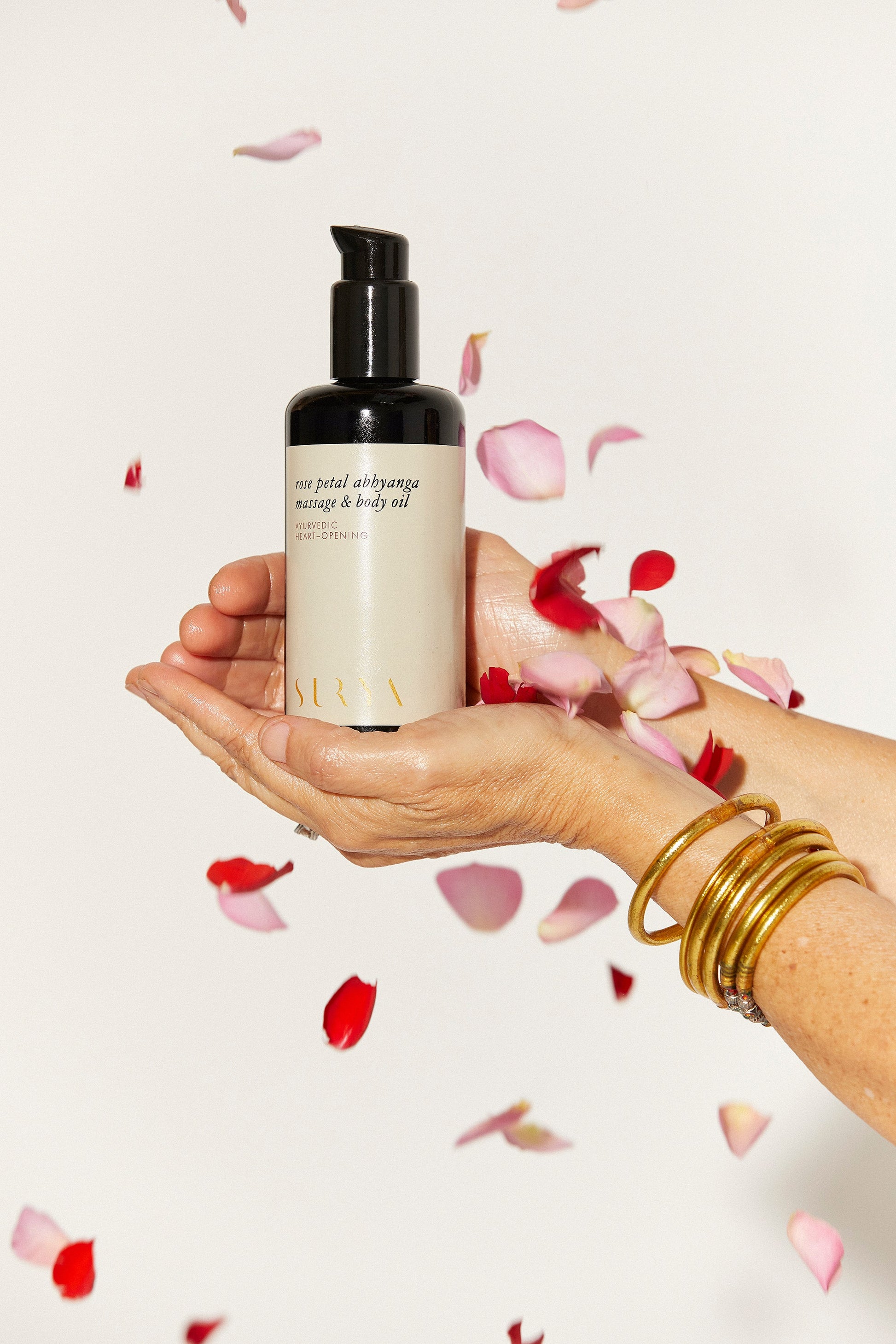A person with gold bangles on their wrist is holding a bottle of Rose Petal Body Oil from Surya, crafted using organic ingredients. The background is white, with rose petals scattered around and gently falling through the air.