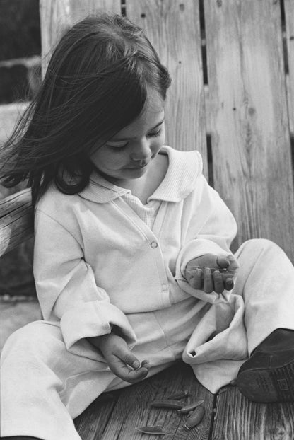 A young child with long hair, dressed in the TAKE CARE TOP hemp by cabane childrenswear, sits thoughtfully on a wooden bench and looks down at small objects in their hand. The photo is black and white.