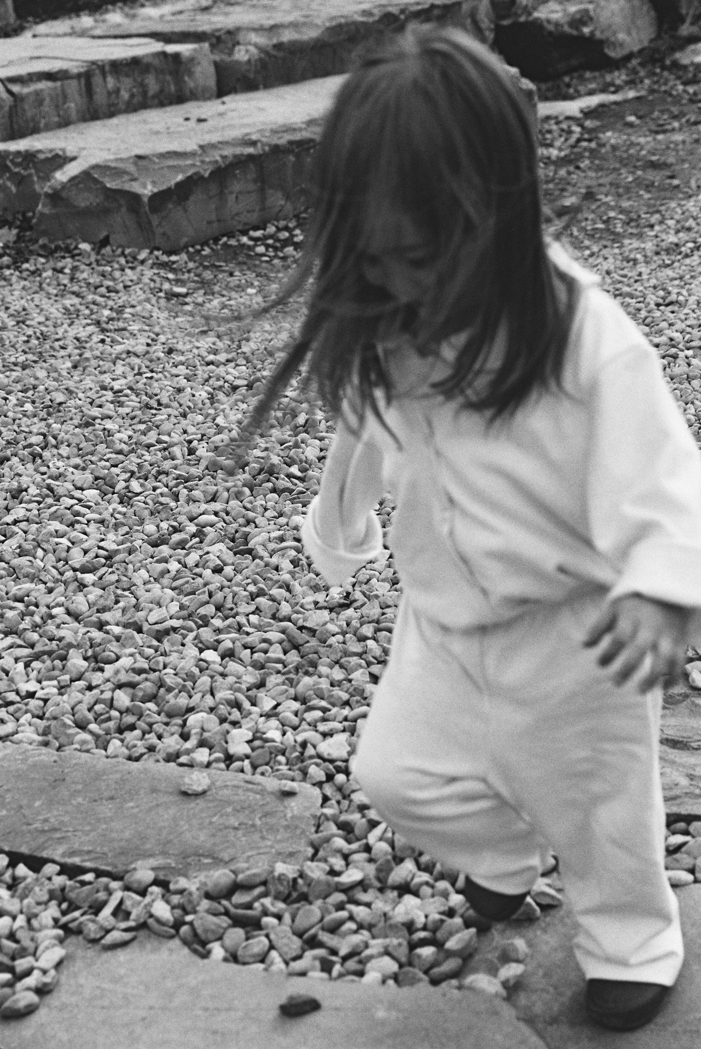 A young child, partly hidden by long hair, walks on a stone path wearing the cabane childrenswear TAKE CARE TOP hemp—ethically handmade from hemp & organic cotton. The black-and-white image highlights the light-colored top amidst small rocks.